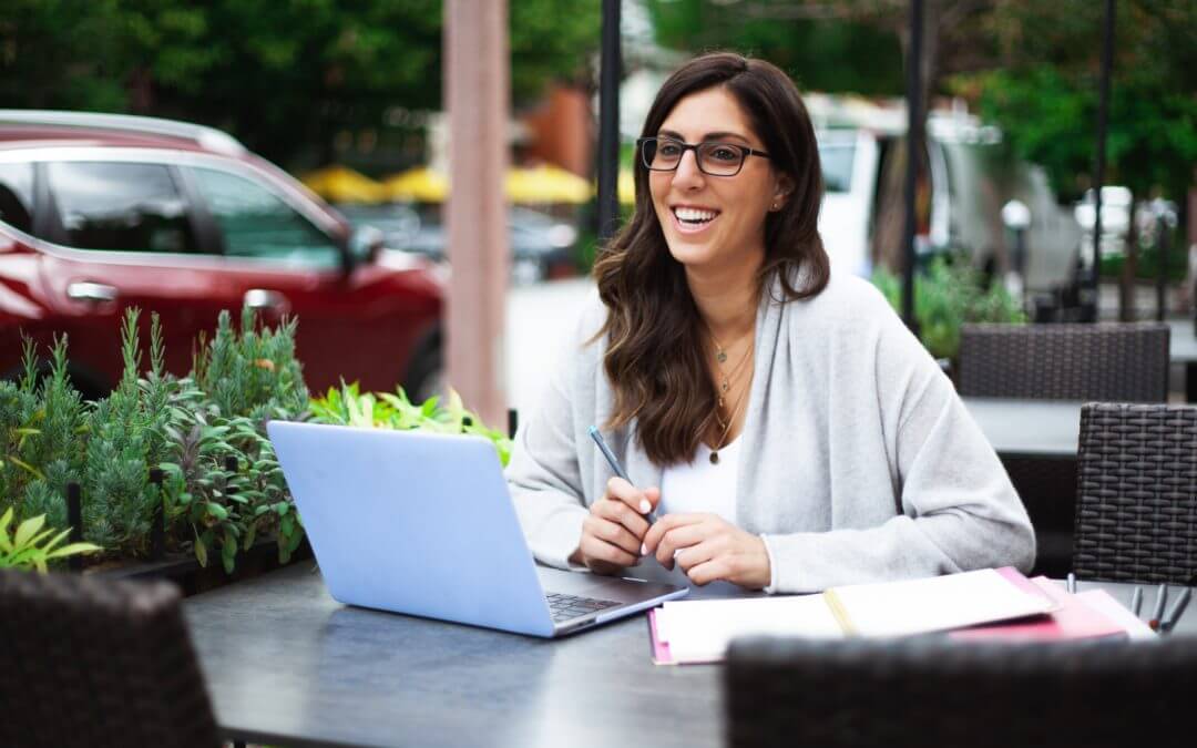 should you go freelance? woman is smiling in front of a laptop in a public place. She has long brown hair and glasses