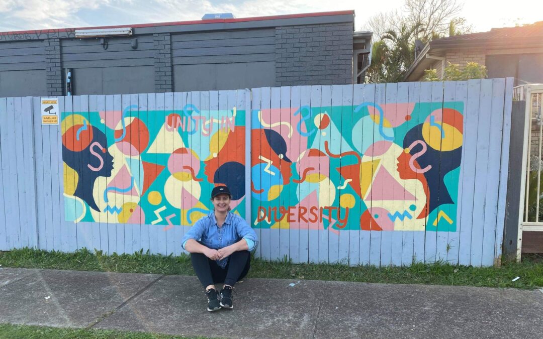 Freelance artists Danielle Said sits in front of a whole fence mural.