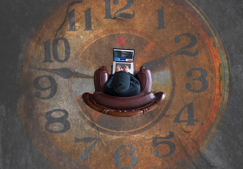 A figure sits in a swivel chair in the middle of a giant floor clock. They are wearing a hoodie and on their laptop. The figure is shot with a birds eye view to donate how to estimate time or time passing in some way.