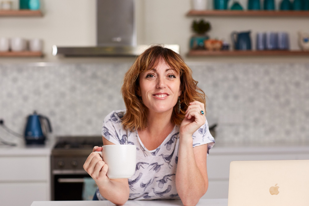 Photo of Kate Toon. She's sitting in a kitchen, holding a coffee mug. Her hair is shoulder length brown and her eyes are brown. She is smiling. Kate recently spoke to us on Crowdcast regarding local SEO.