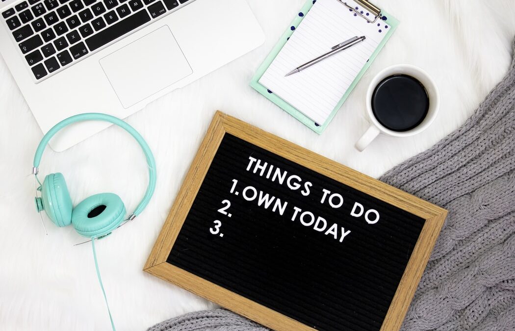A flat lay of a freelance to do list is shot from above. The to do list is a black peg board with white writing that reads things to do 1. own today and 2 and r are blank. There are a coffee, headset and a tablet near by.