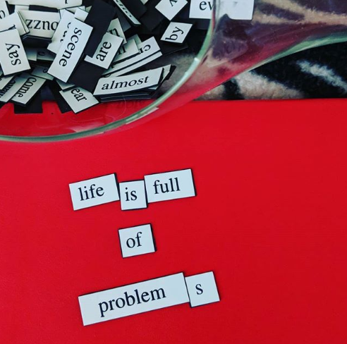 Magnetic words that read "life is full of problems" sit on a red journal. To the top of that is a glass container filled with the rest of the magnetic poetry set. This is used to get people reflecting on freelancing self-care in lockdown now that Melbourne Australia is in stage 4 lockdown.
