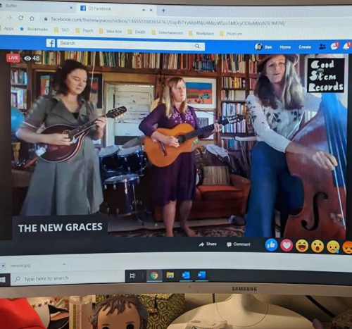 The band the new graces are showing off their virtual creativity by playing online. three women are playing stringed instruments in a living room via a computer monitor.