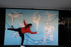 Eva Klusacek. is leaning over her artwork for a local rink. She looks like one of the skaters and she holds her leg high to draw on the wall.