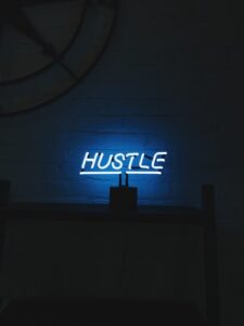 a neon light on a wall reads hustle to demonstrate hustle culture. The room is dark except for the light.