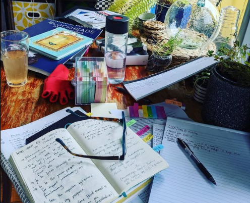 A wooden oval table is covered with journals, books and palm cards as I plan the freelance blogger submission guidelines and program for 2021. There is a water bottle and a tall glass with tepache in it as well as glasses.