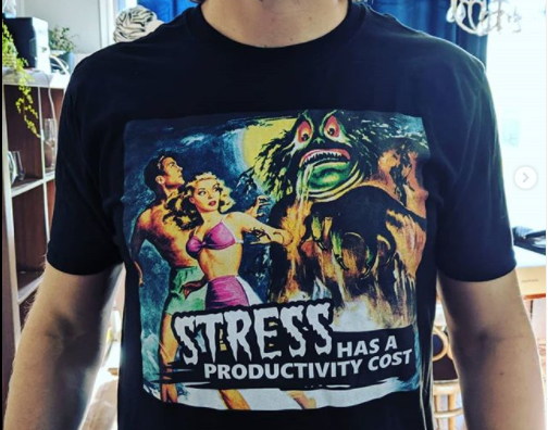 A man stands wearing a tee shirt that reads stress has a productivity cost as the monster of procrastination tries to eat the fair maiden, retro horror movie style. Tee shirt is available on the Freelance Jungle redbubble shop.