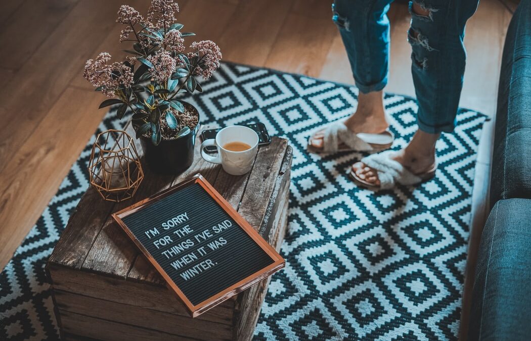 A pegboard sits on a crate coffee table with a drink and a vase of flowers. There are a person's feet in strappy sandals and 3/4 pants nearby. The pegboard reads "i'm sorry for the things I said when it was winter".