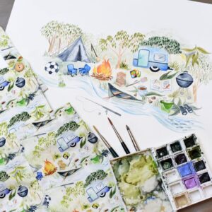 Beautiful hand painted fabric sits next to sketches of the art and a tray of watercolour paint. It reflects a camping scene with bush, triangle tent and round bbq with a blue caravan