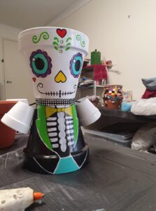 A pot is stuck to another pot to make for a triangular body and head. It is painted into a suit and has big flowers for eyes and an upturned love heart nose. 
