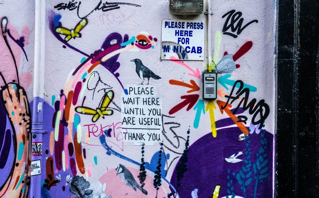 A graffiti covered wall includes a sign that reads "please wait here until you are useful" written in black texta on a white pasted piece of paper