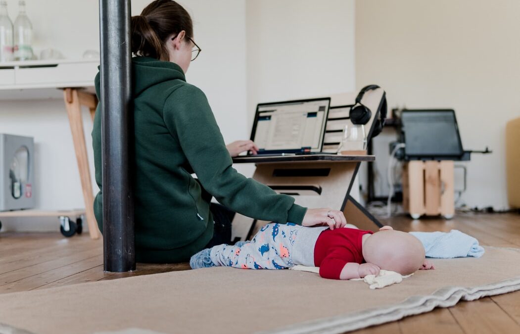 A woman is engaged in remote work on her laptop. She has her back against a pole, sitting on the floor. One hand on laptop, one hand on her baby on a rug beside her