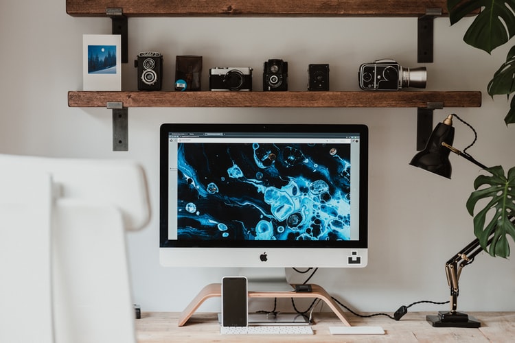 A mac monitor is on a desk. A wooden shelf above shows trinkets and speakers in an ideal telecommuting office setup.