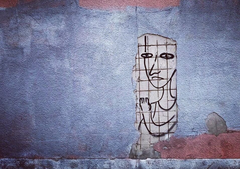 A paste up of a person is stuck to a blue concrete wall. it has a downward mouth and upraised hand.
