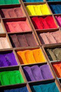 The sun shines on stacked wooden boxes containing powdered pigments in a market in Nepal. The colours range from earthy hues to bright yellows, reds, oranges, purples and greens.