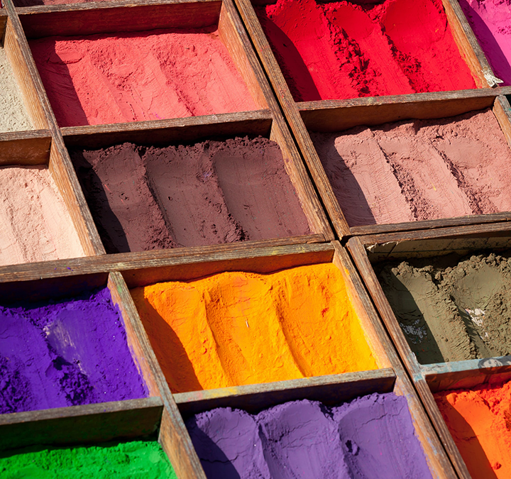 The sun shines on stacked wooden boxes containing powdered pigments in a market in Nepal. The colours range from earthy hues to bright yellows, reds, oranges and purples.