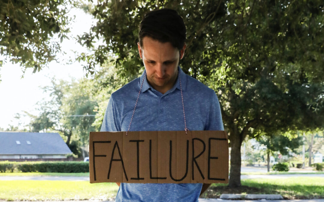 A man stands with the word failure on a sign around their neck with their head hung in creative shame.